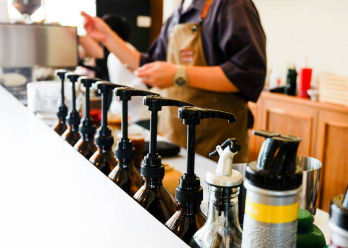 A barista behind the counter at a cafe, taking an order from behind a variety of coffee syrup pumps.